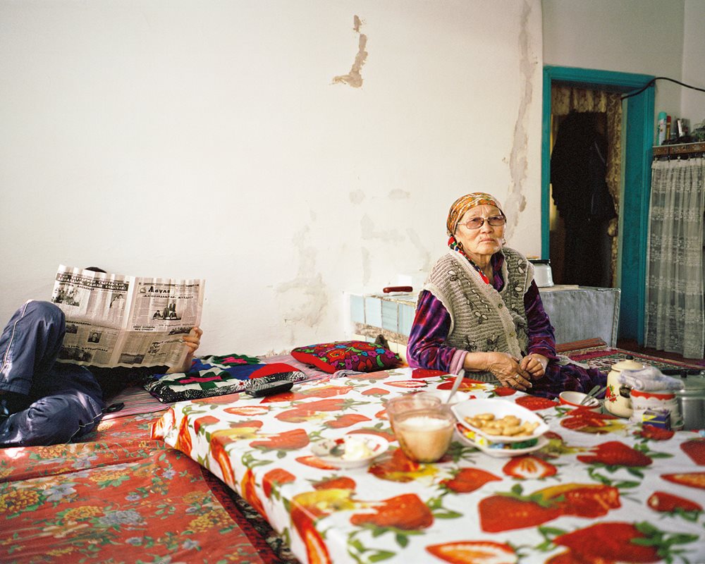 <p>Oltinay watches television while her son reads the paper at home in Aral. She remembers her late husband&rsquo;s job at Aral&rsquo;s fish-processing plant, which has recently started up again.</p>
