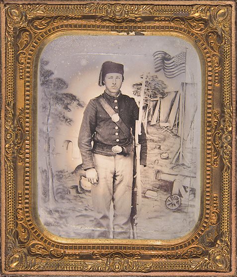 <p>Outfitted with a Zouave fez and equipped with an 1862 Zouave-style sword bayonet, this unidentified Union soldier posed for a portrait against a backdrop depicting a Union military camp</p>
