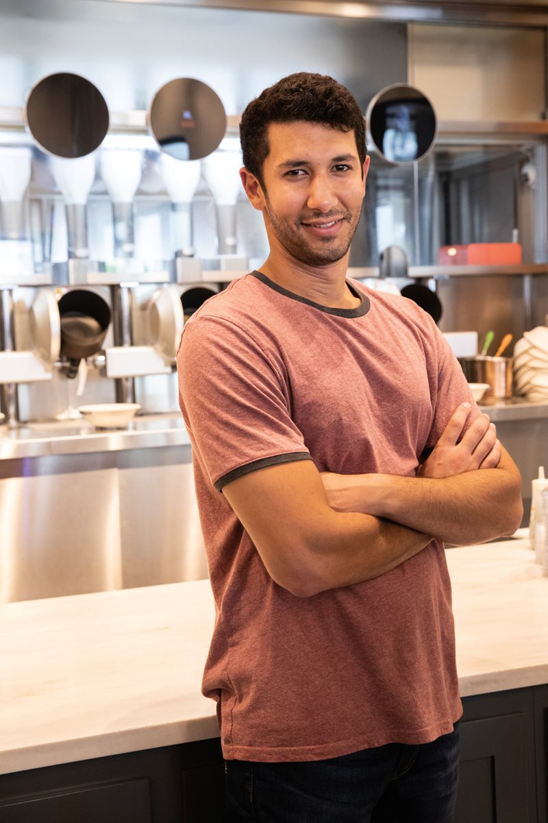 Cofounder of Spyce Michael Farid, 28, stands in front of the array of robotic woks that turned an idea into a thriving business.