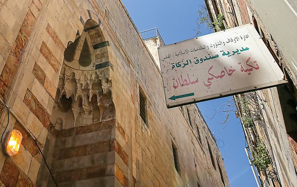 In a narrow street of Old Jerusalem, a sign in Arabic points the way to <em>Takiyah Khaski Sultan</em>, the charitable soup kitchen founded in 1552 by the wife of Ottoman Sultan Suleiman. <strong>Below</strong>&nbsp;Recipients line up outside the courtyard that with the kitchen, a school and a mosque forms the fully endowed charitable complex.